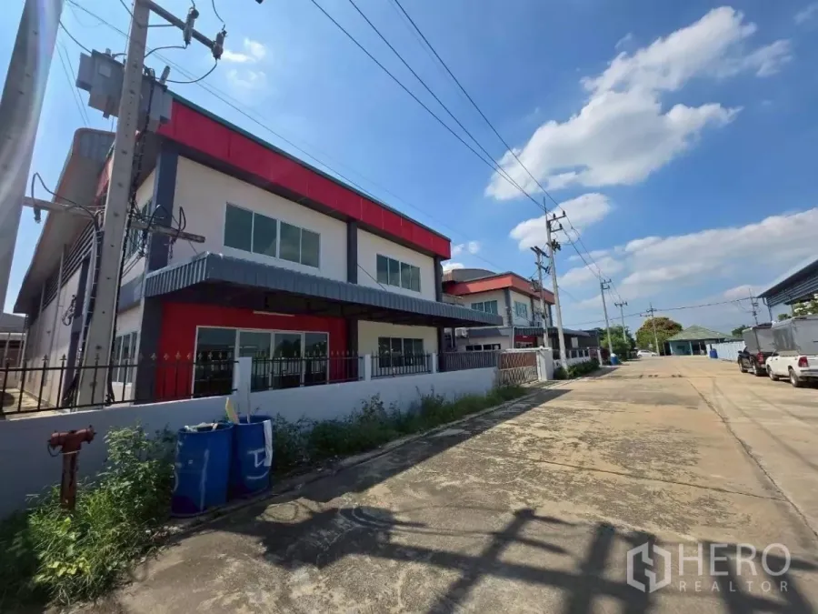 Factory for rent or sale in Mueang Samut Sakhon, Samut Sakhon - Street view of a modern two-storey factory with red accents, fenced perimeter and a wide access road in Samut Sakhon.