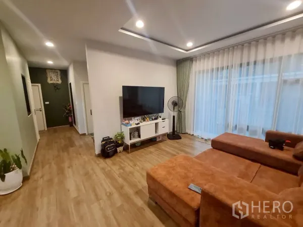 Detached House for rent or sale in Lam Luk Ka, Pathum Thani - Family lounge with an L-shaped sofa, TV console and full-height sheer curtains.