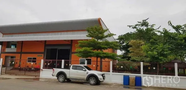 Factory for sale in Bang Phli, Samut Prakan - Street view of an orange warehouse building with gated fence, trees and a pickup truck at the frontage.