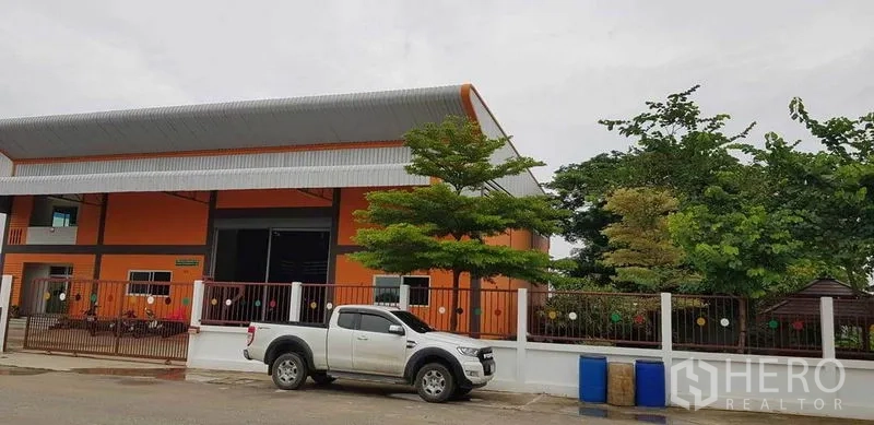 Factory for sale in Bang Phli, Samut Prakan - Street view of an orange warehouse building with gated fence, trees and a pickup truck at the frontage.