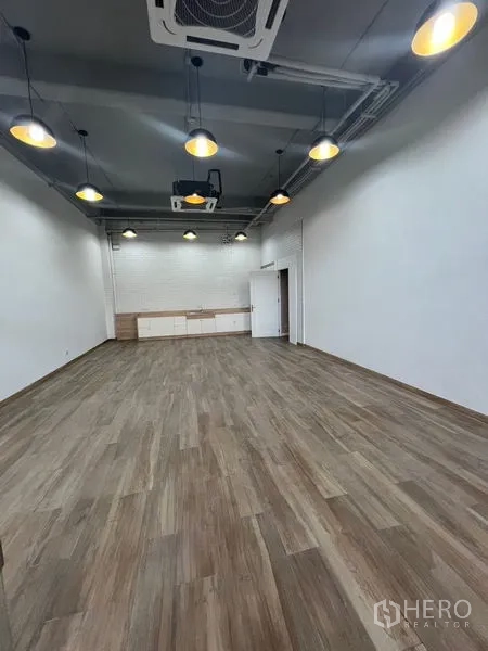 Retail Space for rent - Open‑plan retail interior with high ceiling, pendant lights, wood‑look floors and a rear storage counter.