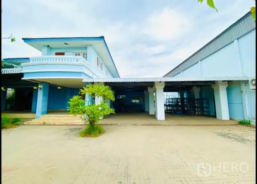 Factory for rent in Sam Phran, Nakhon Pathom - Front view of a light-blue two‑storey office attached to a warehouse with a wide paved yard.