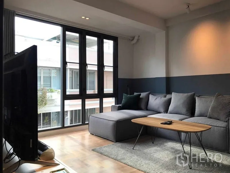 Townhouse for rent or sale in Khlong Toei, Bangkok - Sunlit sitting room with wall‑to‑wall windows, gray sofa and wood floors.