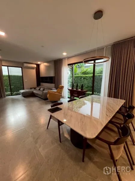 Detached House for rent in Samut Prakan - Open-plan living and dining area with marble table, pendant light, and full-height glass to the garden.