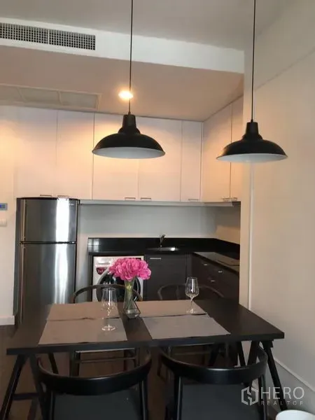 Condominium for rent in Pathum Wan, Bangkok - Modern kitchen with white cabinets, black countertops, stainless refrigerator, and a dining table under pendant lights.