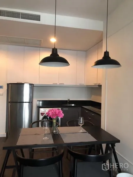 Condominium for rent in Pathum Wan, Bangkok - Modern kitchen with white cabinets, black countertops, stainless refrigerator, and a dining table under pendant lights.