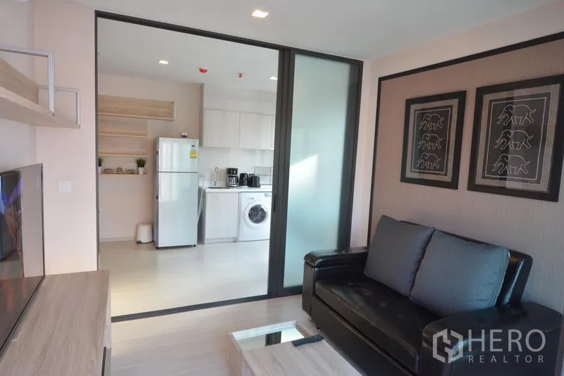 Condominium for sale in Khlong Toei, Bangkok - Bright living room with black sofa, TV console and sliding glass door to a modern kitchen with fridge and washing machine.