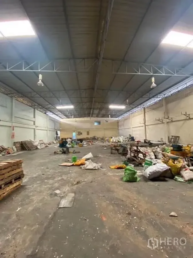 Factory for rent in Samut Sakhon - Wide-span factory hall with steel truss roof, skylights, industrial lights and a concrete floor with materials stored across the area.