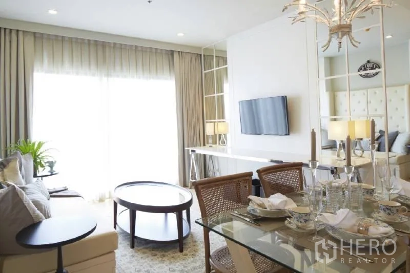 Condominium for rent in Bangkok - Living area facing large windows with TV wall and mirrored panels beside dining space.