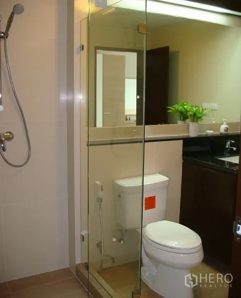 Apartment for rent in Khlong Toei, Bangkok - Modern bathroom with glass-enclosed shower, toilet, and vanity with mirror and dark countertop.