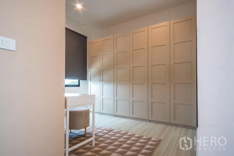 Townhouse for rent or sale in Suan Luang, Bangkok - Dressing room with built-in beige wardrobes, a vanity desk, and a window with blackout blind.