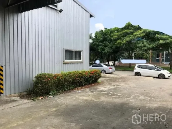 Warehouse for rent in Pak Kret, Nonthaburi - Side view of warehouse with small windows, shrub landscaping and open parking area beside tree-lined road.