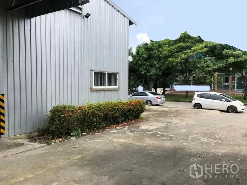 Warehouse for rent in Pak Kret, Nonthaburi - Side view of warehouse with small windows, shrub landscaping and open parking area beside tree-lined road.