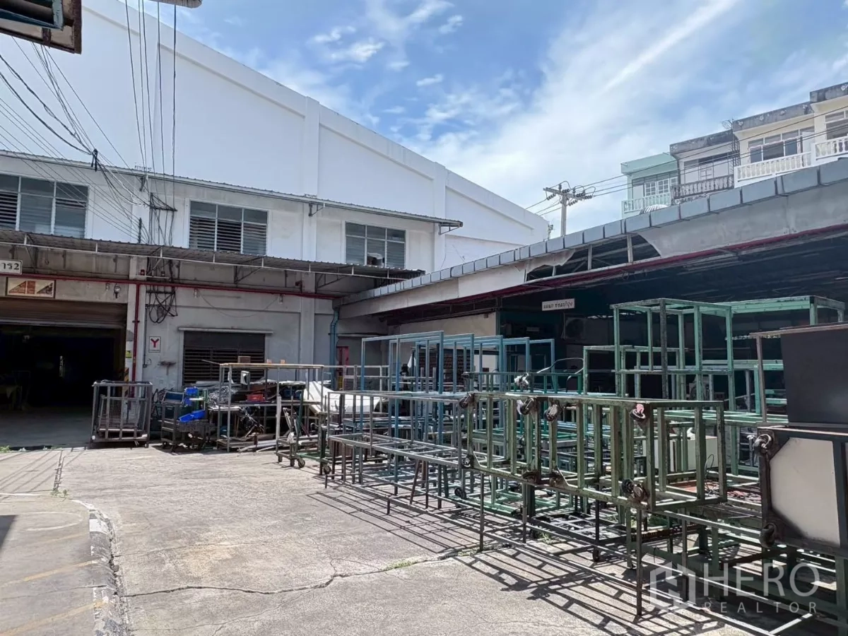 Factory for rent or sale in Bang Khun Thian, Bangkok - Open yard with metal storage racks and adjacent workshop buildings on a concrete surface.