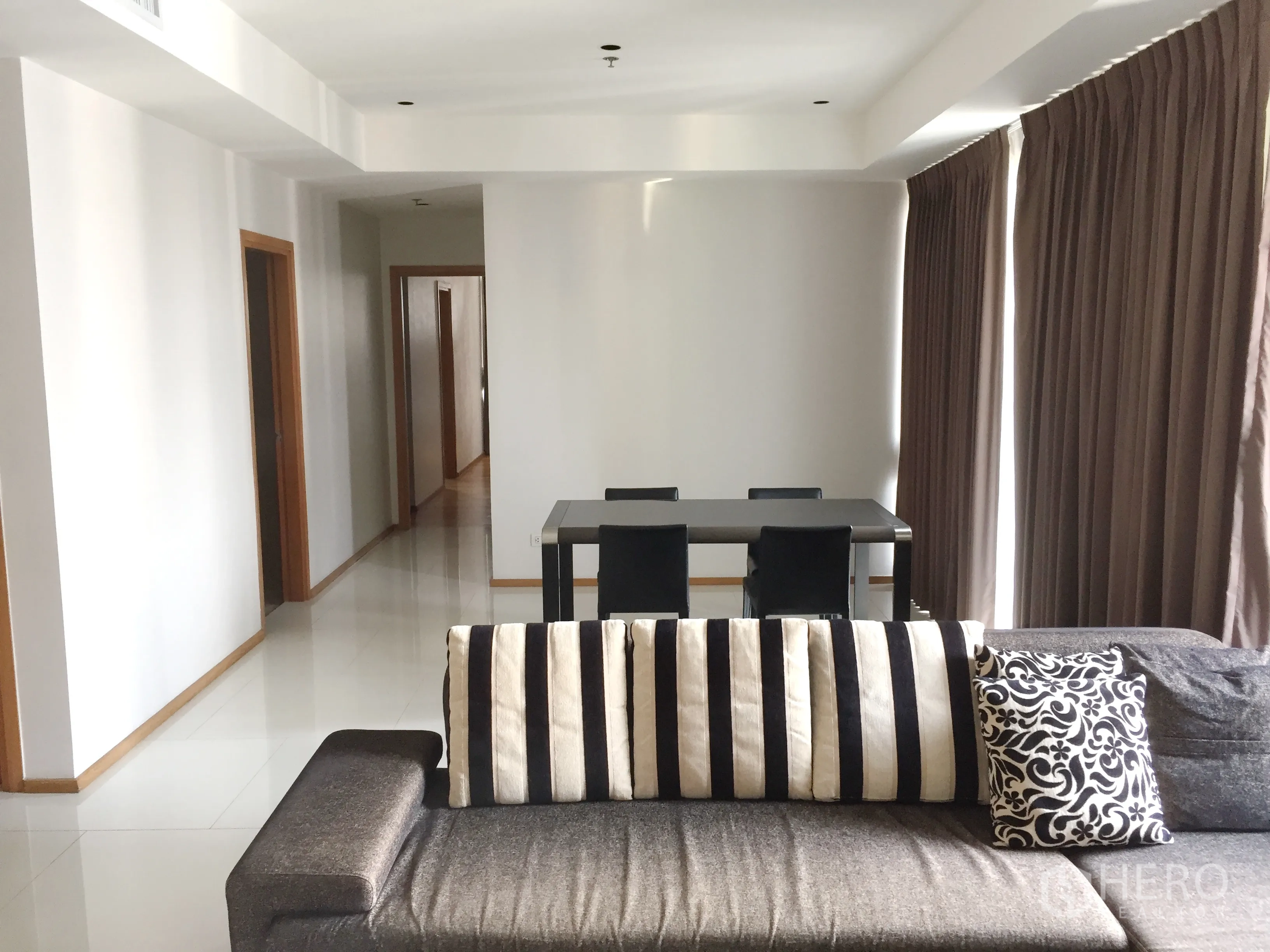 Condominium for rent in Khlong Toei, Bangkok - Spacious lounge with striped-cushion sofa, dining table for four, and hallway leading to bedrooms.
