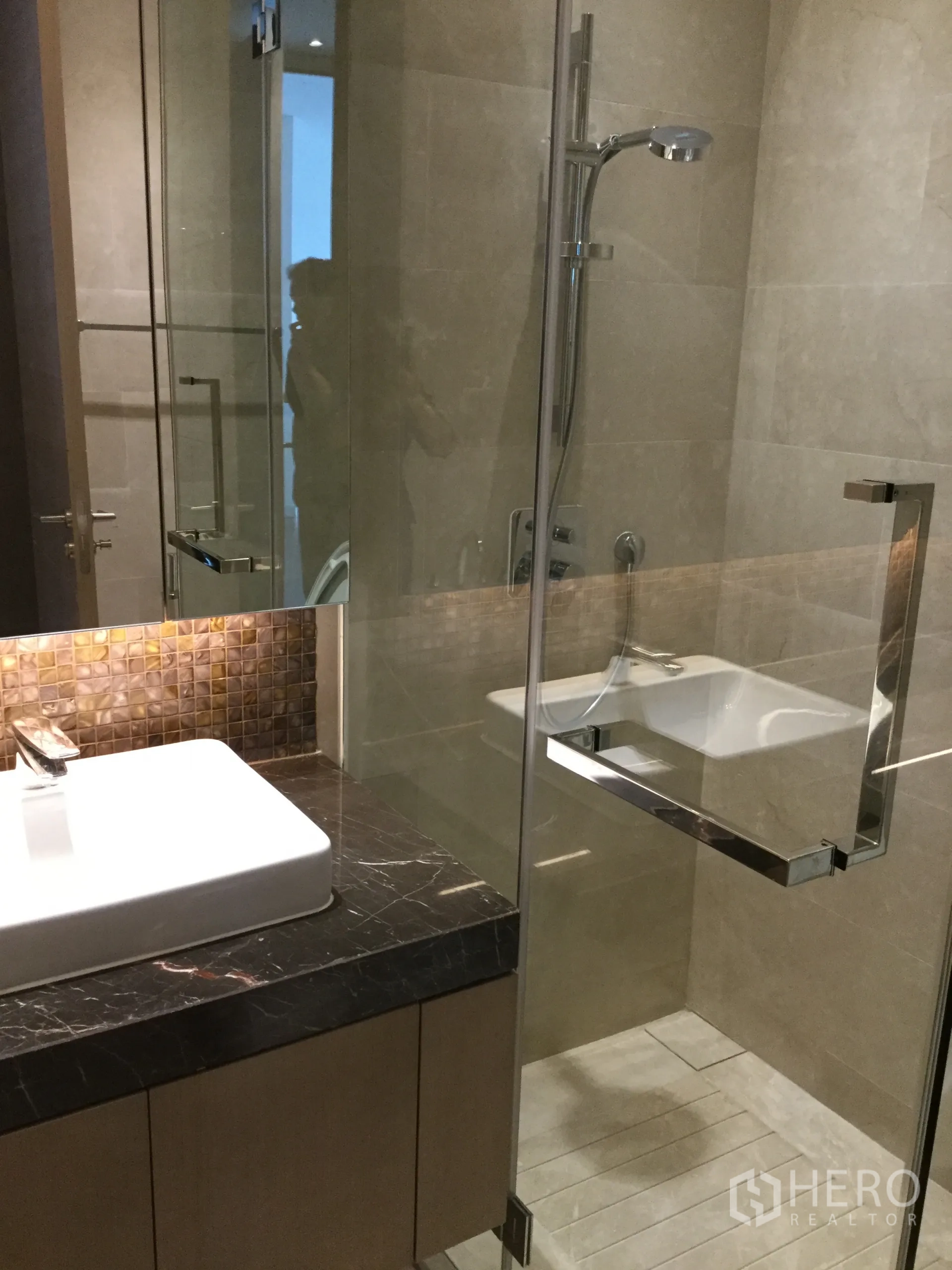 Condominium for rent in Pathum Wan, Bangkok - Hotel-style bathroom with glass shower enclosure, square basin on dark marble counter and beige tiles.