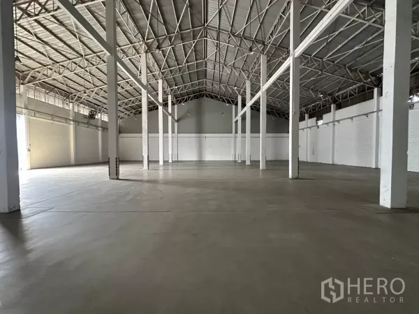 Warehouse for rent in Phra Khanong, Bangkok - Wide storage area with concrete flooring, white pillars and a roller shutter door at the far end.