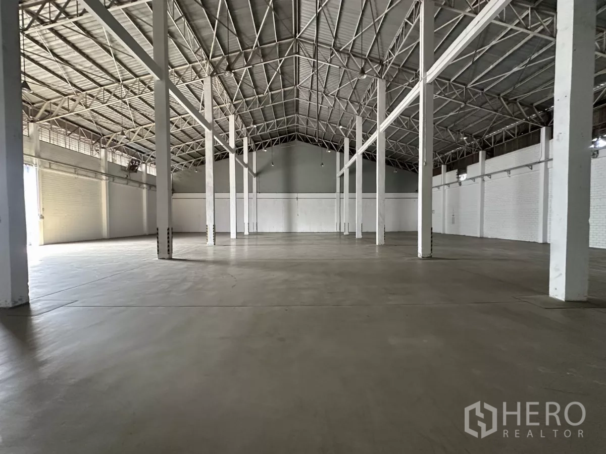 Warehouse for rent in Phra Khanong, Bangkok - Wide storage area with concrete flooring, white pillars and a roller shutter door at the far end.