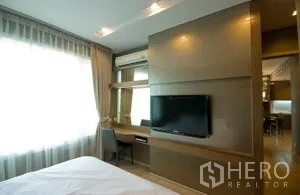 Condominium for rent in Khlong Toei, Bangkok - Bedroom feature wall with mounted TV, study desk, and natural light from a wide window.