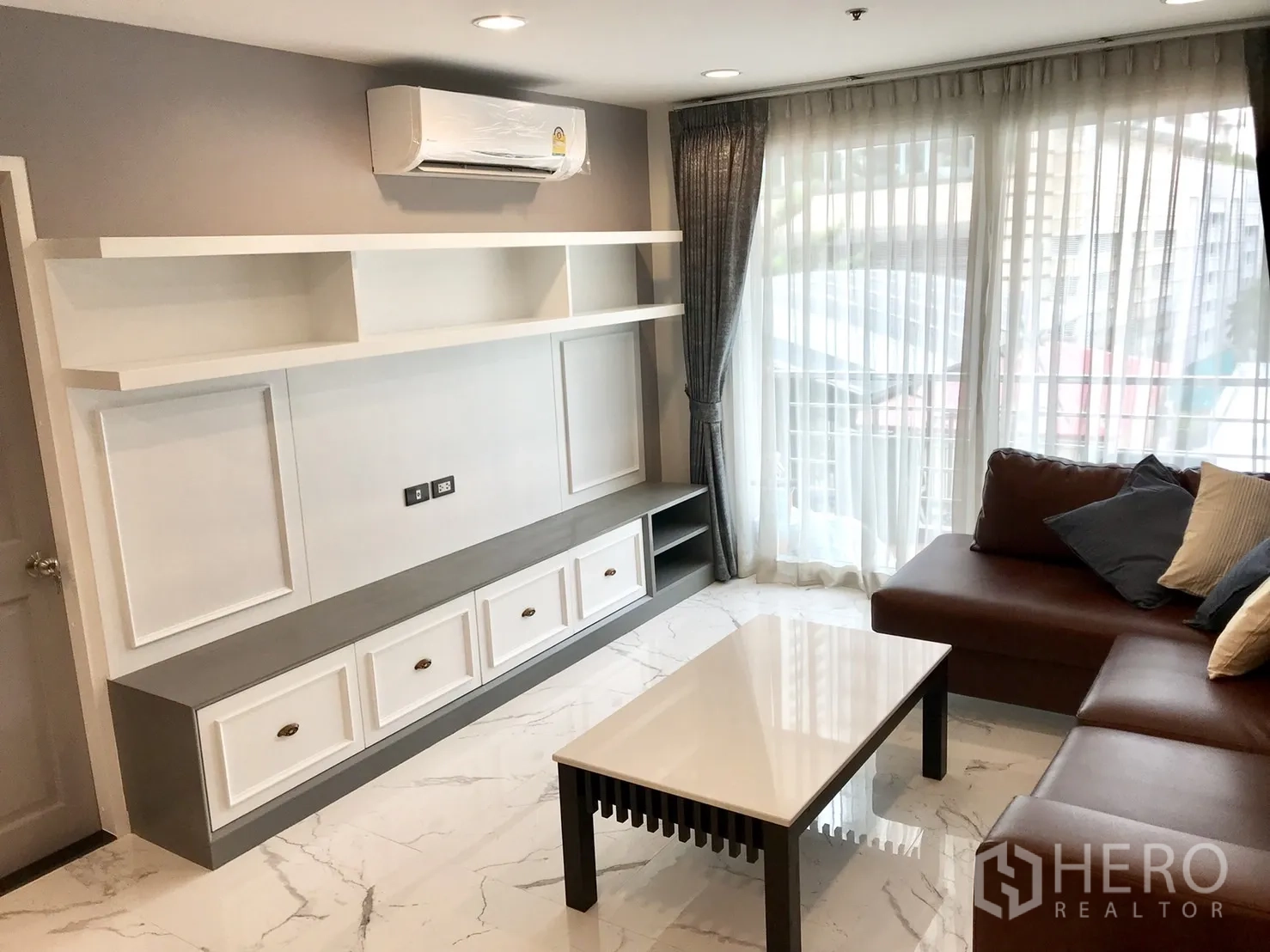 Condominium for rent or sale in Khlong Toei, Bangkok - Built-in TV console with open shelves and air-conditioning beside a sofa and floor-to-ceiling windows.