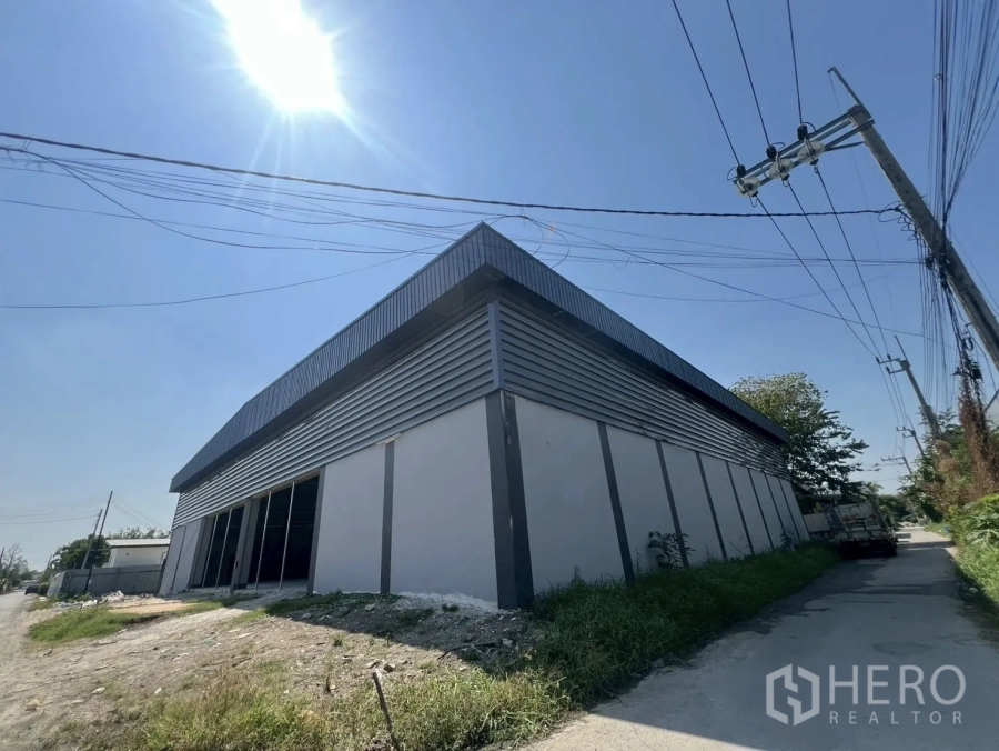 Warehouse for rent in Bueng Kum, Bangkok - Corner exterior of a modern steel-clad warehouse with wide door openings and roadside access under a bright sunny sky.