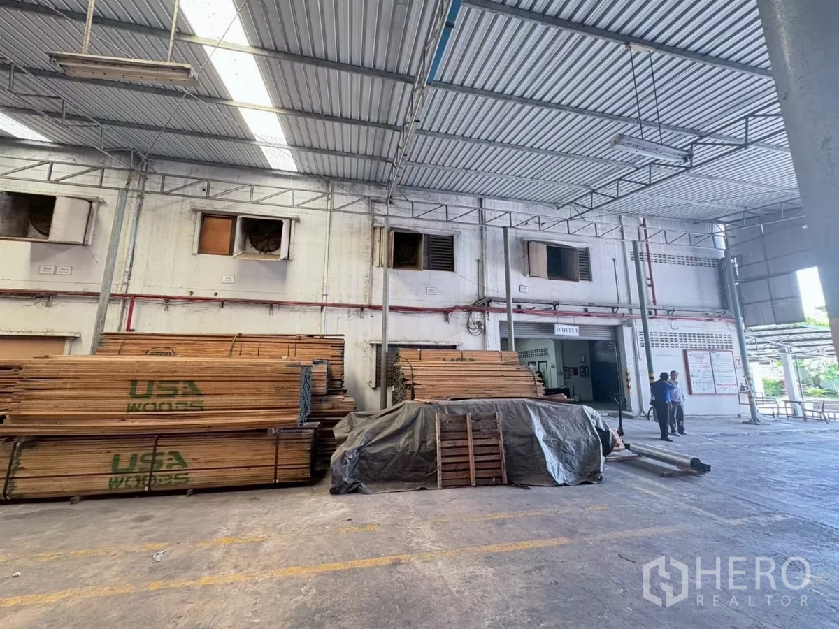 Factory for rent or sale in Bang Khun Thian, Bangkok - Stacked timber under a covered loading area beside the factory facade.