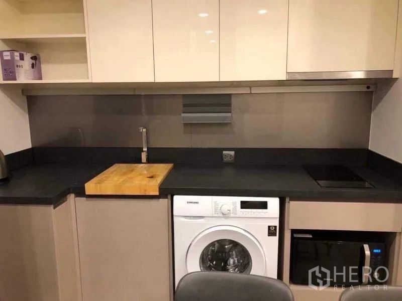Condominium for rent in Khlong Toei, Bangkok - Modern kitchenette with dark countertop, induction hob, sink, upper cabinets, and front-load washer.