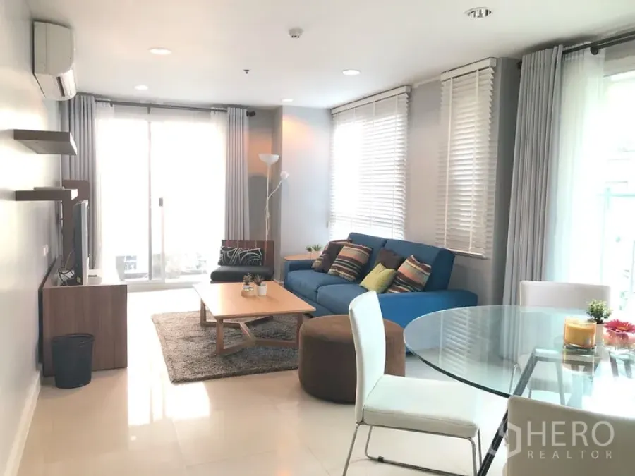Condominium for rent or sale in Khlong Toei, Bangkok - Bright living room with blue sofa, glass dining set and sliding doors to a balcony.
