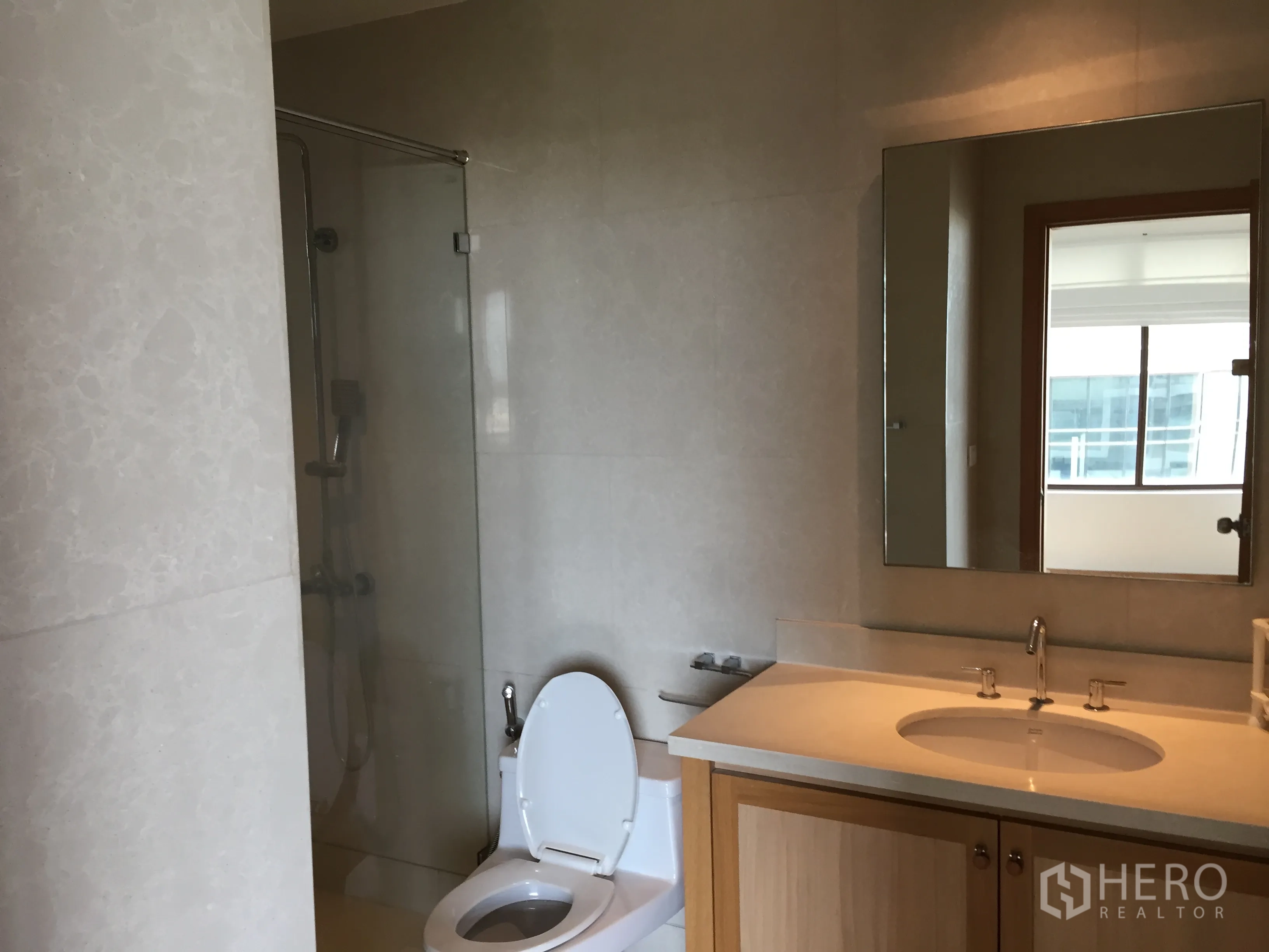 Condominium for rent in Khlong Toei, Bangkok - Modern bathroom featuring glass-enclosed shower, wide vanity with mirror and neutral stone tiles.