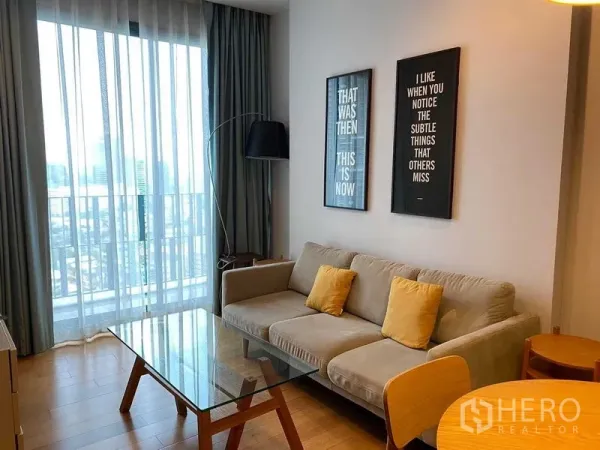 Condominium for rent in Khlong Toei, Bangkok - Living room with gray sofa, glass coffee table and floor-to-ceiling window showing city view.