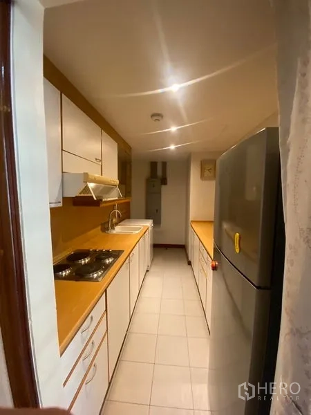 Condominium for rent in Pathum Wan, Bangkok - Galley kitchen with white cabinets, cooktop, sink, range hood, and a full-size refrigerator.