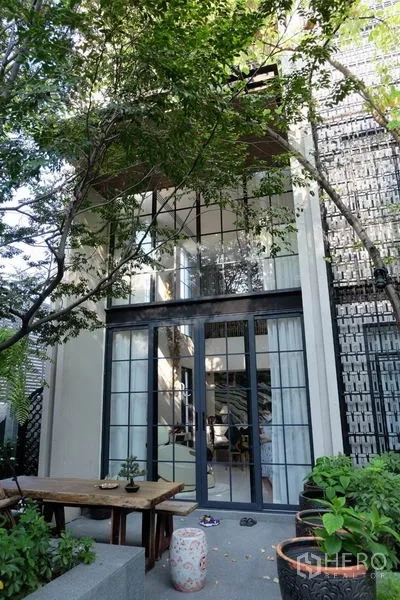 Townhouse for rent in Khlong Toei, Bangkok - Garden patio with trees facing a double‑height townhouse facade and black‑framed glass doors.