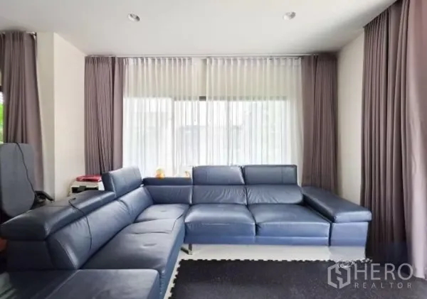 Detached House for sale in Bang Kapi, Bangkok - Living area corner featuring a large blue L-shaped leather sofa and layered curtains.
