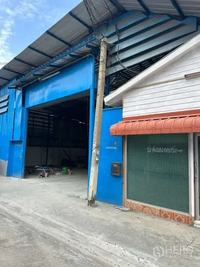 Warehouse for rent in Prawet, Bangkok - Blue metal warehouse exterior with a wide roller-shutter entry beside a small office on a narrow concrete lane in Prawet, Bangkok.