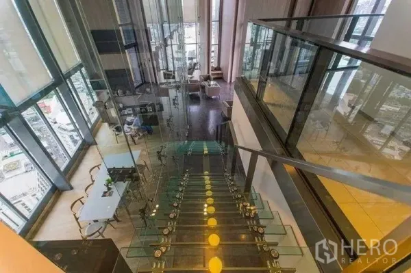 Condominium for rent in Khlong Toei, Bangkok - Glass staircase leading to a double-height lounge with floor-to-ceiling windows and Bangkok city views.