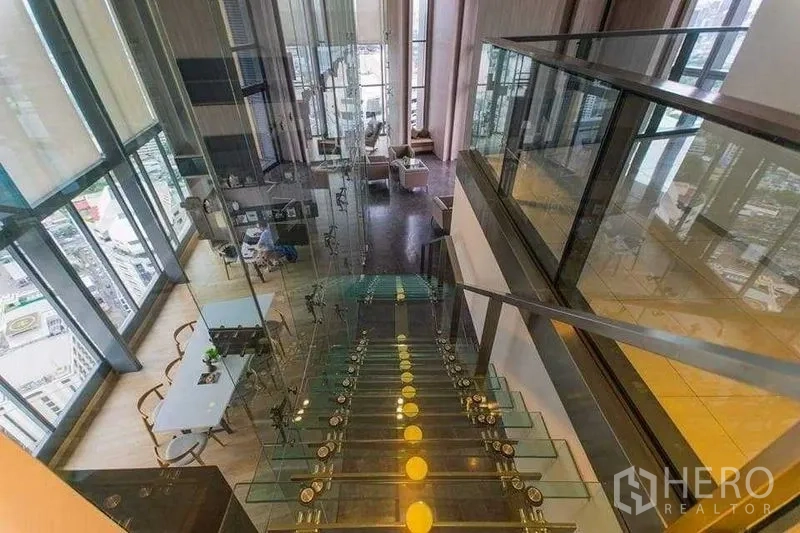 Condominium for rent in Khlong Toei, Bangkok - Glass staircase leading to a double-height lounge with floor-to-ceiling windows and Bangkok city views.