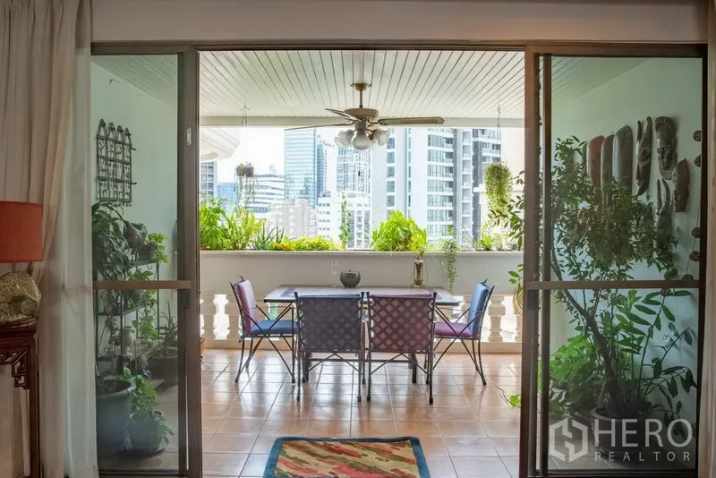 Condominium for rent in Khlong Toei, Bangkok - Covered balcony with sliding glass doors, ceiling fan, potted greenery and skyline view set for alfresco dining.