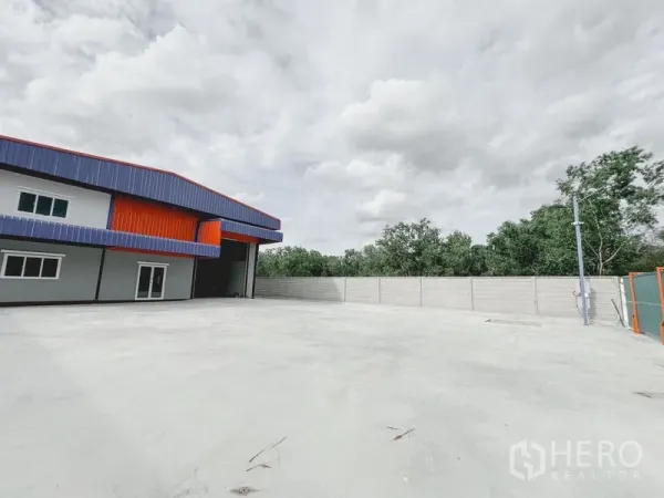 Warehouse for rent or sale in Khlong Luang, Pathum Thani - Expansive concrete yard beside the warehouse with a tall roller shutter, perimeter wall and trees beyond.