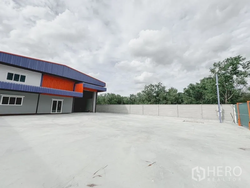 Warehouse for rent or sale in Khlong Luang, Pathum Thani - Expansive concrete yard beside the warehouse with a tall roller shutter, perimeter wall and trees beyond.