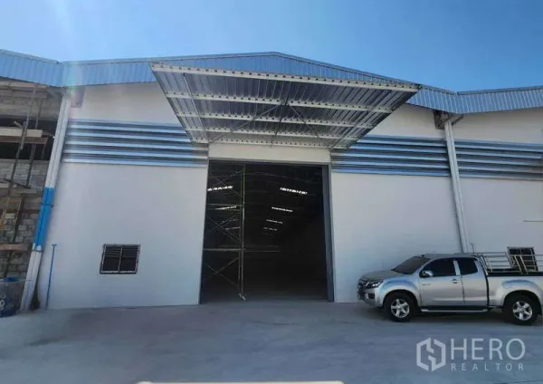 Warehouse for rent in Sam Phran, Nakhon Pathom - Front facade of modern warehouse showing metal awning and truck‑friendly driveway under a clear blue sky.