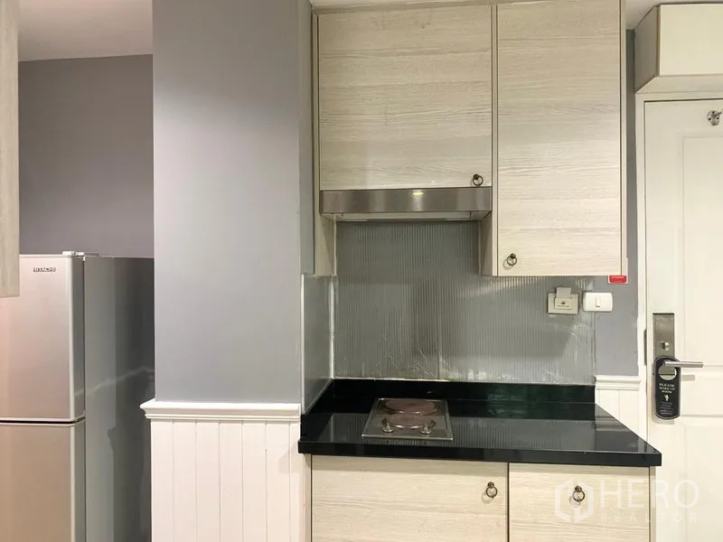 Condominium for rent in Khlong Toei, Bangkok - Compact kitchenette with cooktop, hood, black countertop and fridge in the condo.