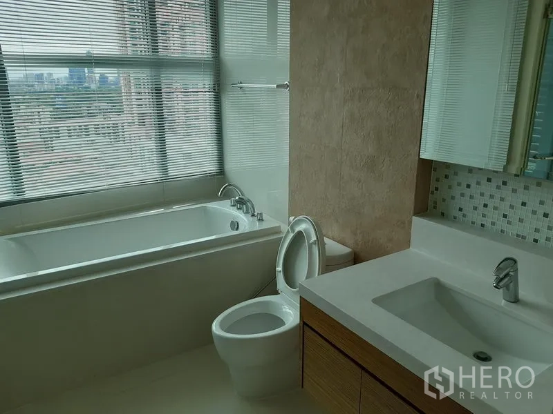Condominium for rent or sale in Khlong Toei, Bangkok - Bathroom with bathtub beneath a wide window, toilet and long vanity with basin.