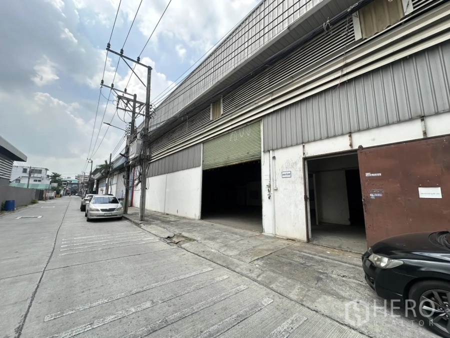 Warehouse for rent in Suan Luang, Bangkok - Street view of an industrial warehouse with a green roller shutter and side entrance along a concrete lane.