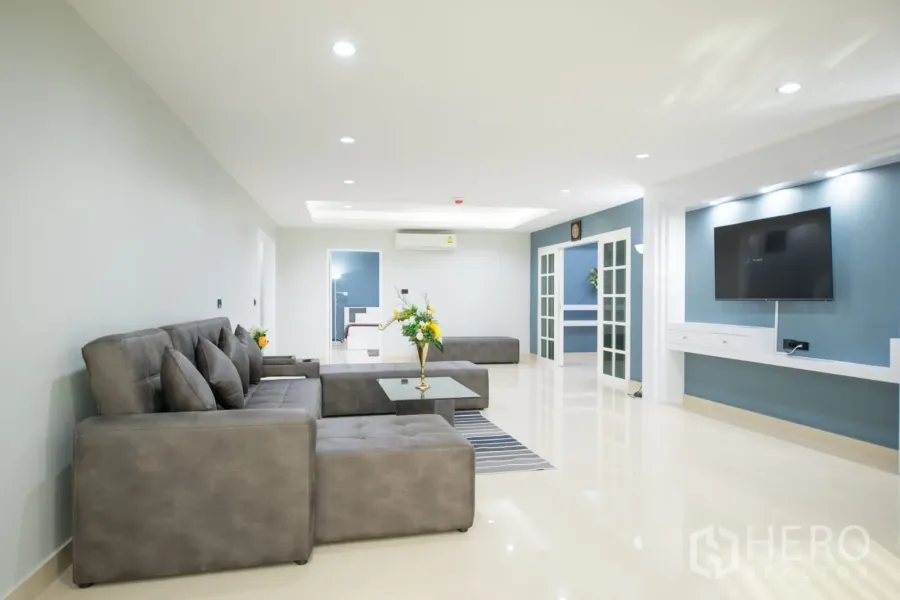 Condominium for rent or sale in Khlong Toei, Bangkok - Spacious living room in a Bangkok condo with grey sectional sofa, built‑in TV wall, recessed lighting and glass doors.