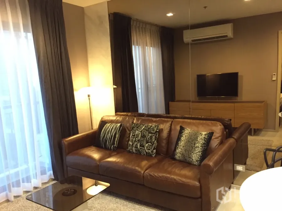 Condominium for rent in Khlong Toei, Bangkok - Warm living room with brown leather sofa, floor lamp, TV console and large curtained windows.