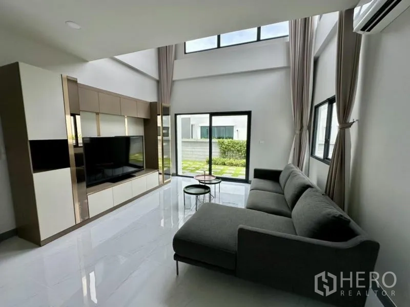 Detached House for rent in Bang Phli, Samut Prakan - Living room with L-shaped sofa, tall curtains, and sliding doors to the yard.