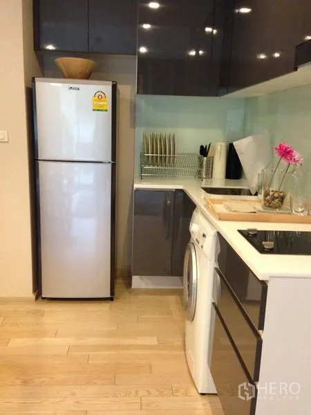 Condominium for rent in Watthana, Bangkok - Compact L-shaped kitchen with glass backsplash, refrigerator, cooktop and front-load washer.