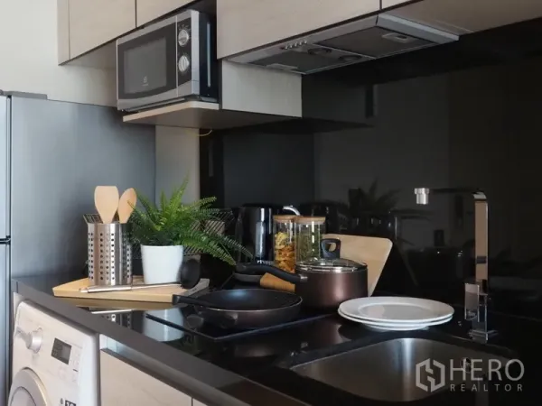 Condominium for sale in Khlong Toei, Bangkok - Compact modern kitchenette with black countertop, sink, hob, microwave, washing machine and cookware.