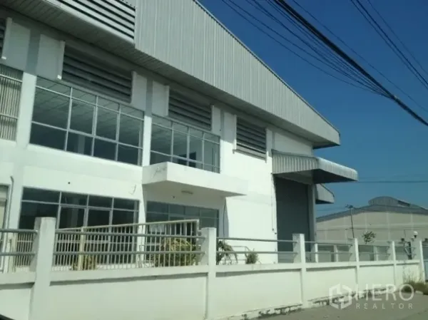 Warehouse for rent in Samut Prakan - Street-side view of a fenced warehouse with large windows and a wide roller shutter door in Samut Prakan.