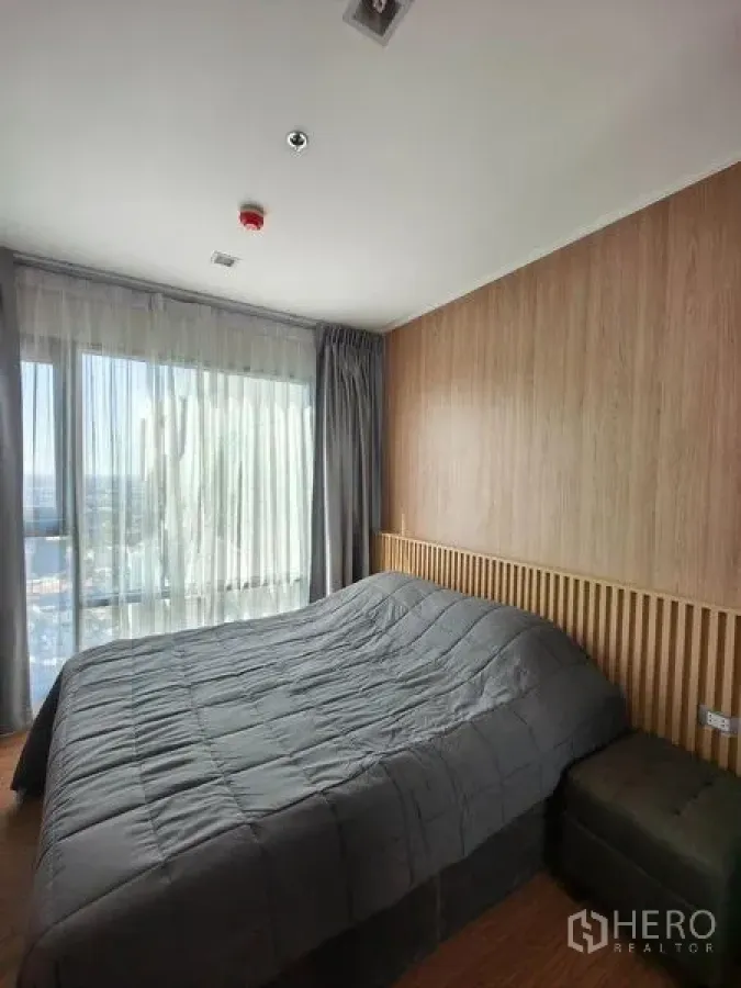 Condominium for rent in Khlong Toei, Bangkok - Bright bedroom with floor-to-ceiling window, gray bedding, wood accent wall and city view.